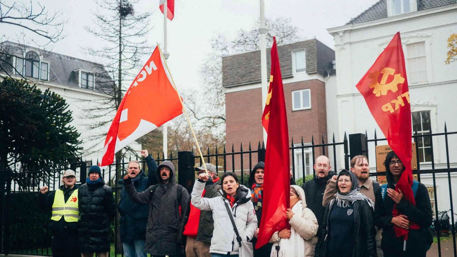 Demonstranten staan met een vuist omhoog voor de Ambassade van Polen in Den Haag. Ze hebben vlaggen van de NCPN en TKP vast.