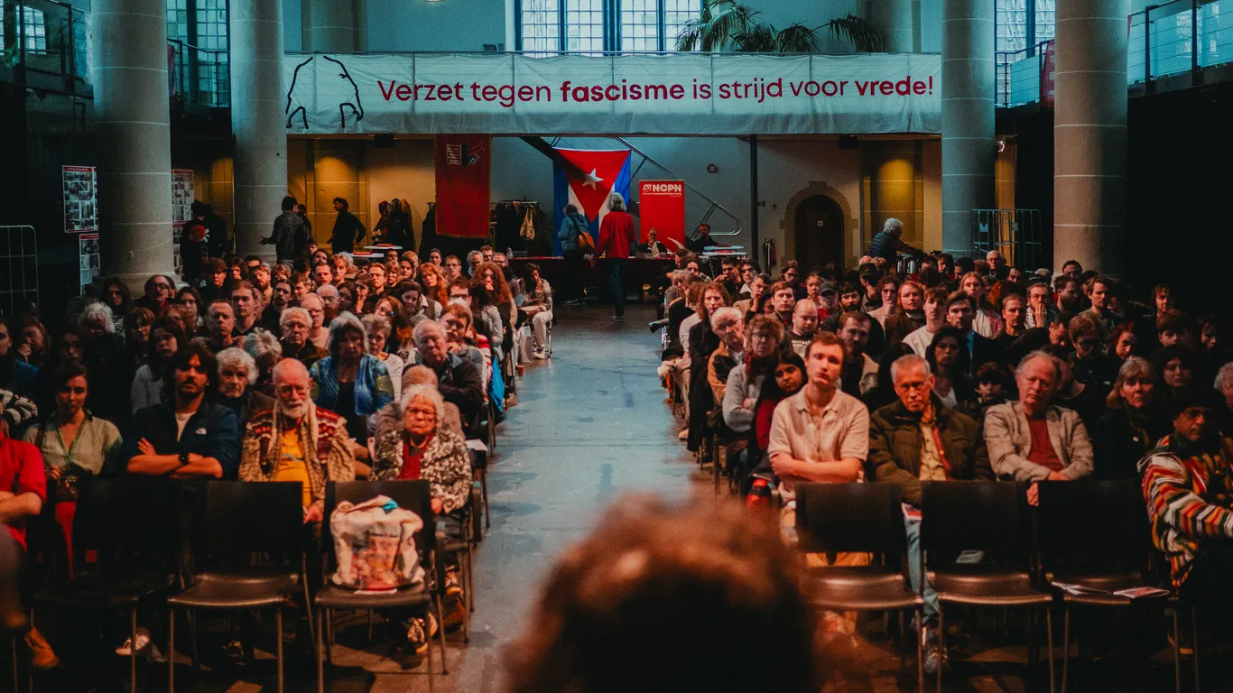 Aanwezigen luisteren aandachtig naar een panelgesprek, dat plaatsvindt als onderdeel van de Februaristakingherdenking in de Zuiderkerk. Achterin hangt een spandoek waarop staat "Verzet tegen fascisme is strijd voor vrede!"