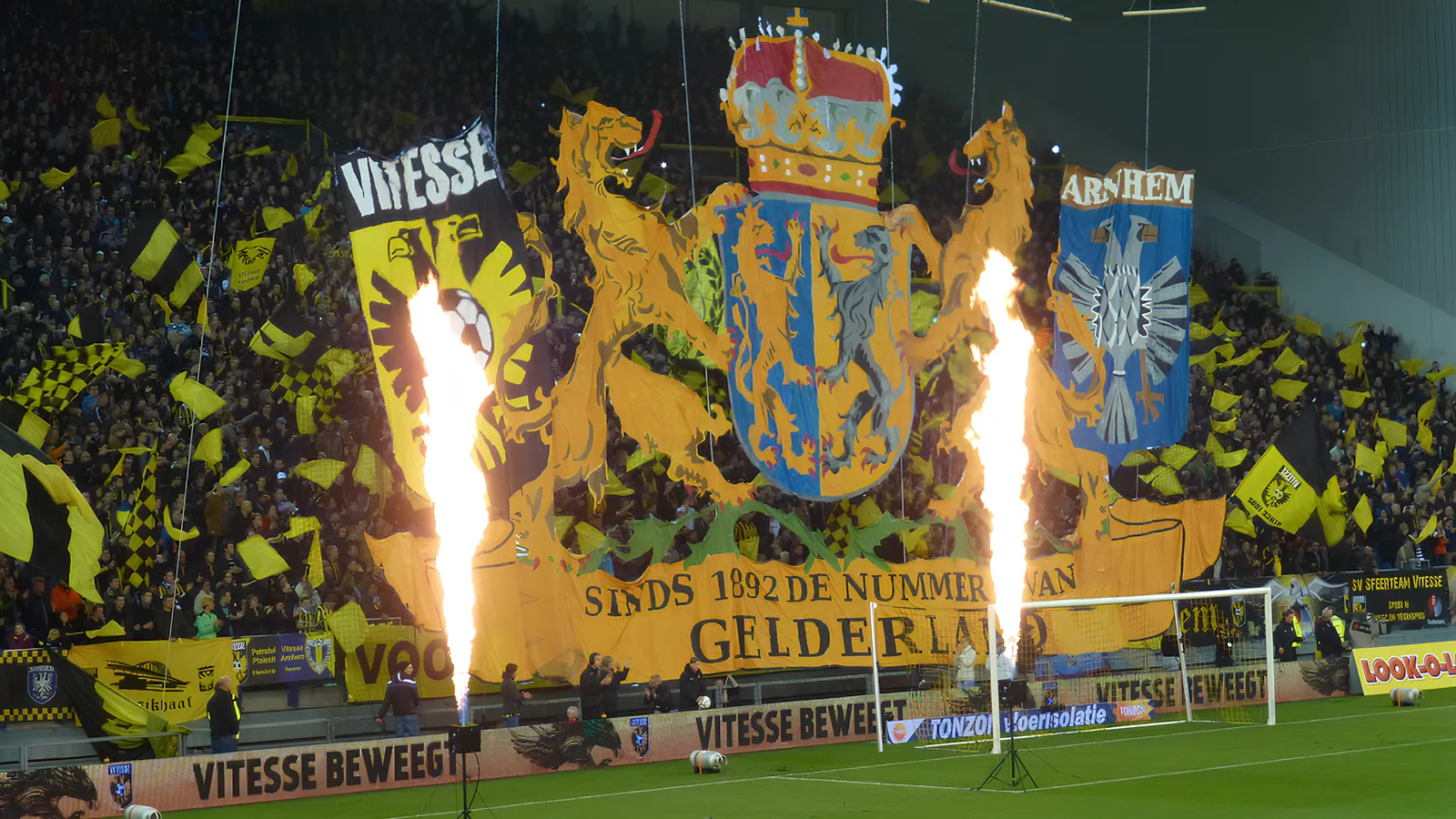 Vitesse-fans wapperen met zwart-gele vlaggen op de tribune van het Gelredome. Vooraan hangt een enorme banier met daarop de vlaggen van Vitesse en Arnhem, en in het midden het wapen van Gelderland. Daaronder staat "Sinds 1892 de nummer 1 van Gelderland"
