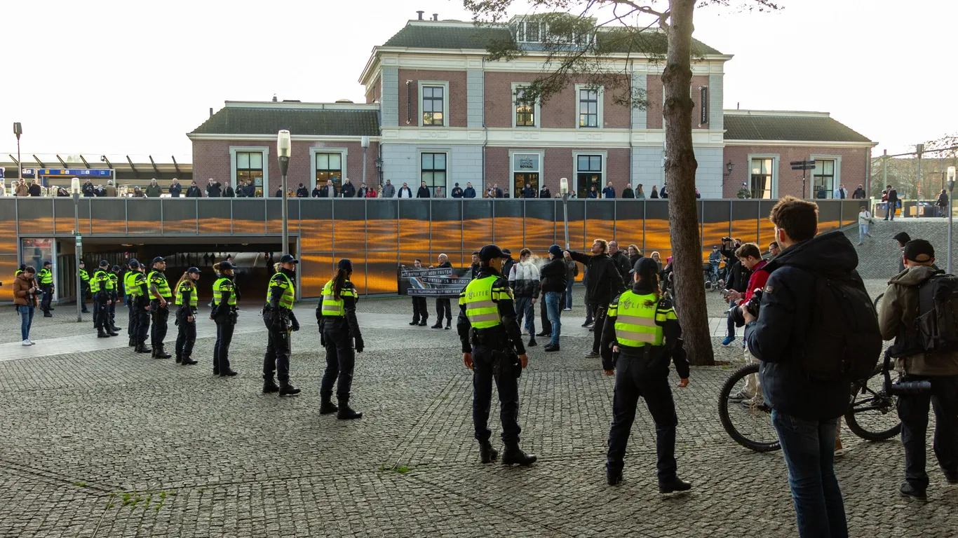 Neonazi's staan voor het station van Apeldoorn, met een politielinie die ze scheidt van de veel grotere tegendemonstratie. Op een spandoek staat "Het systeem kent geen fouten, het systeem is de fout. Gooi de volksverraders uit Den Haag!"