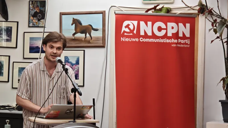 Hidde spreekt de conferentie toe van achter een spreekstoel, naast hem een vlag van de NCPN.