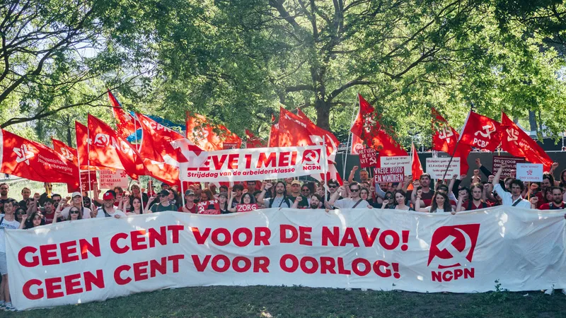 Een grote groep mensen leden en sympathisanten van de NCPN en CJB staan achter een banner, met daarop "Geen cent voor de NAVO! Geen cent voor oorlog!". Daarboven rode vlaggen en een banner die leest "Leve 1 mei: strijddag van de arbeidersklasse"