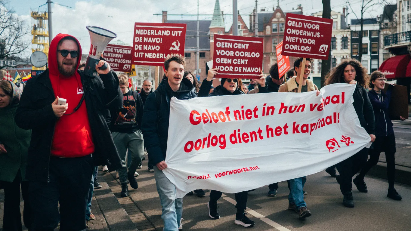 Mensen lopen in een protestmars door Den Haag. Iemand met een megafoon loopt voorop, er worden leuzen geroepen. Vooraan een banner waarop staat "Geloof niet in hun verhaal, oorlog dient het kapitaal"