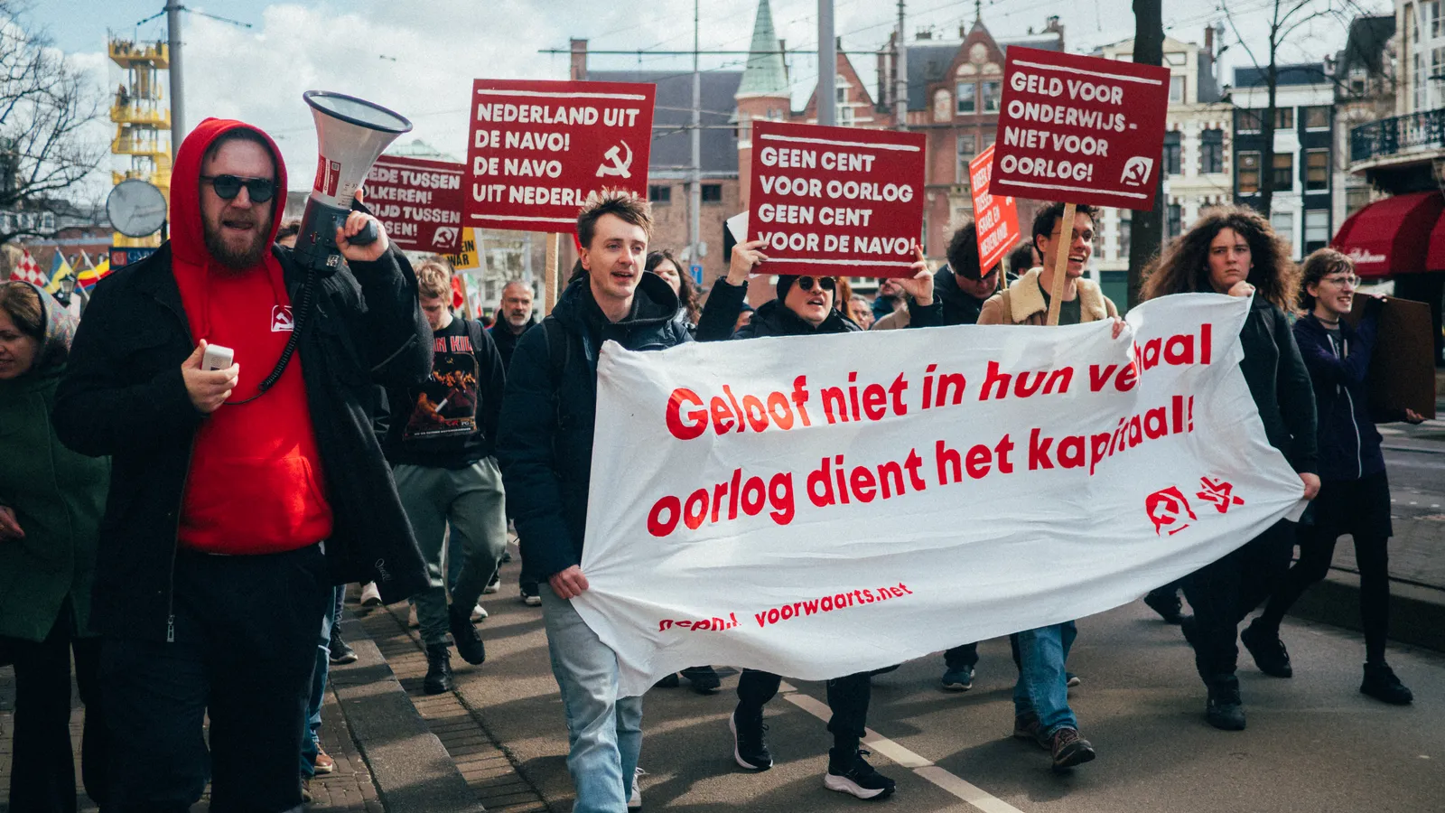 Mensen lopen in een protestmars door Den Haag. Iemand met een megafoon loopt voorop, er worden leuzen geroepen. Vooraan een banner waarop staat "Geloof niet in hun verhaal, oorlog dient het kapitaal"