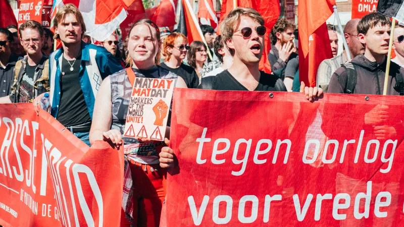 Op de 1 mei-mars in Amsterdam staan mensen achter een banner waarvan zichtbaar is "Tegen oorlog, voor vrede". Daarachter iemand met een bordje waarop staat "May day, may day: workers of the world unite!"