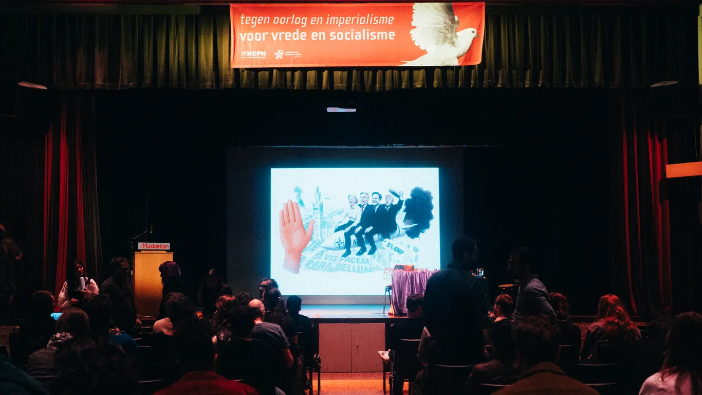 Bezoekers gaan zitten voor een nog leeg podium, met daarboven een banner met daarop een duif en daarnaast "Tegen oorlog en imperialisme - voor vrede en socialisme"