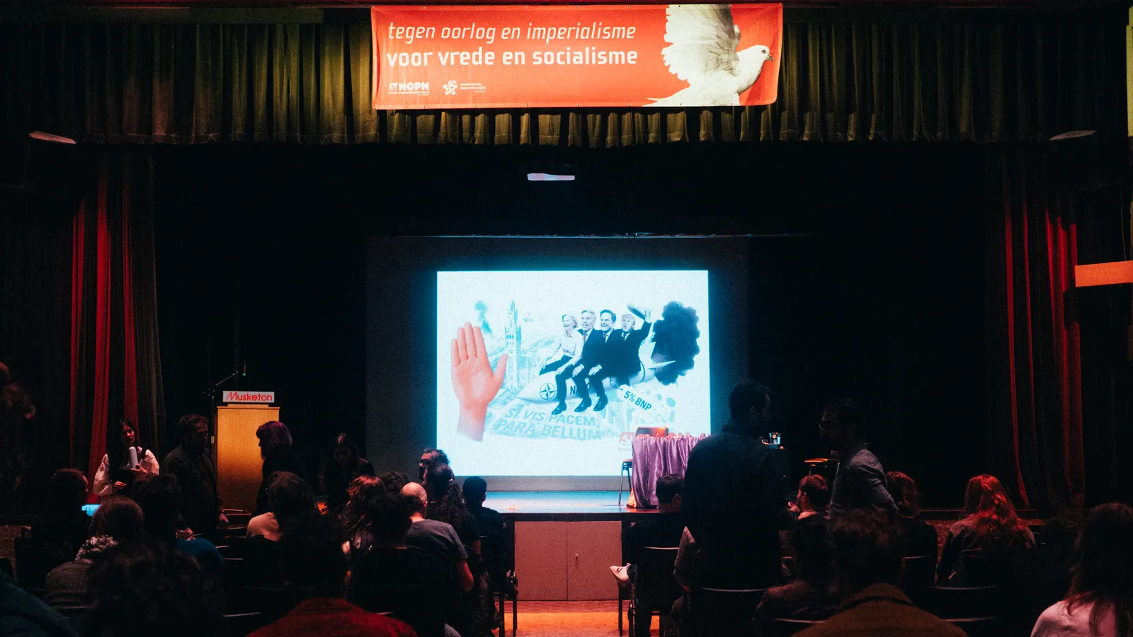 Bezoekers gaan zitten voor een nog leeg podium, met daarboven een banner met daarop een duif en daarnaast "Tegen oorlog en imperialisme - voor vrede en socialisme"