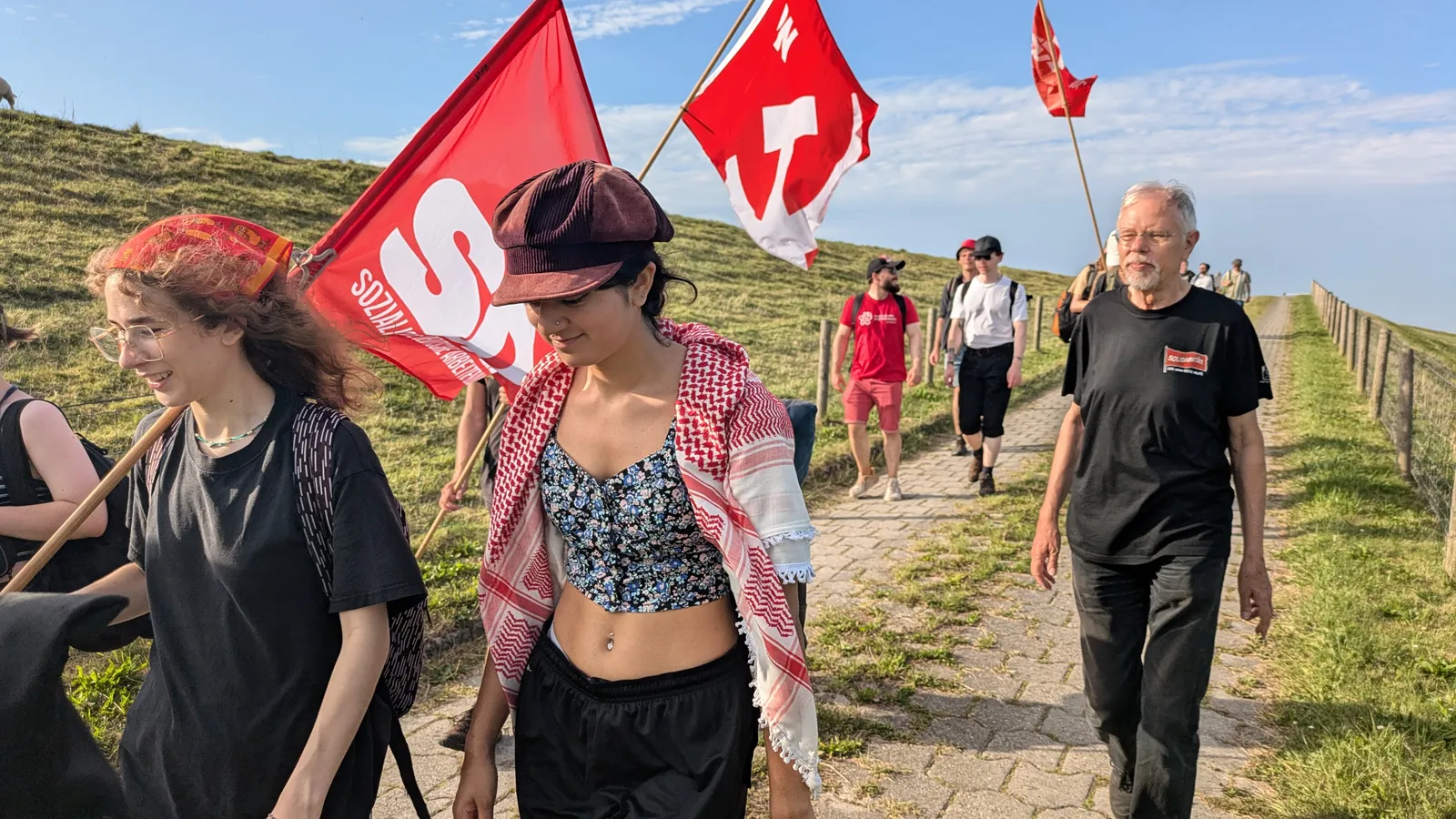 Mensen lopen over het platteland van Groningen, langs een historische vluchtroute van de Rode Hulp. Ze dragen vlaggen van de NCPN, CJB en SDAJ.