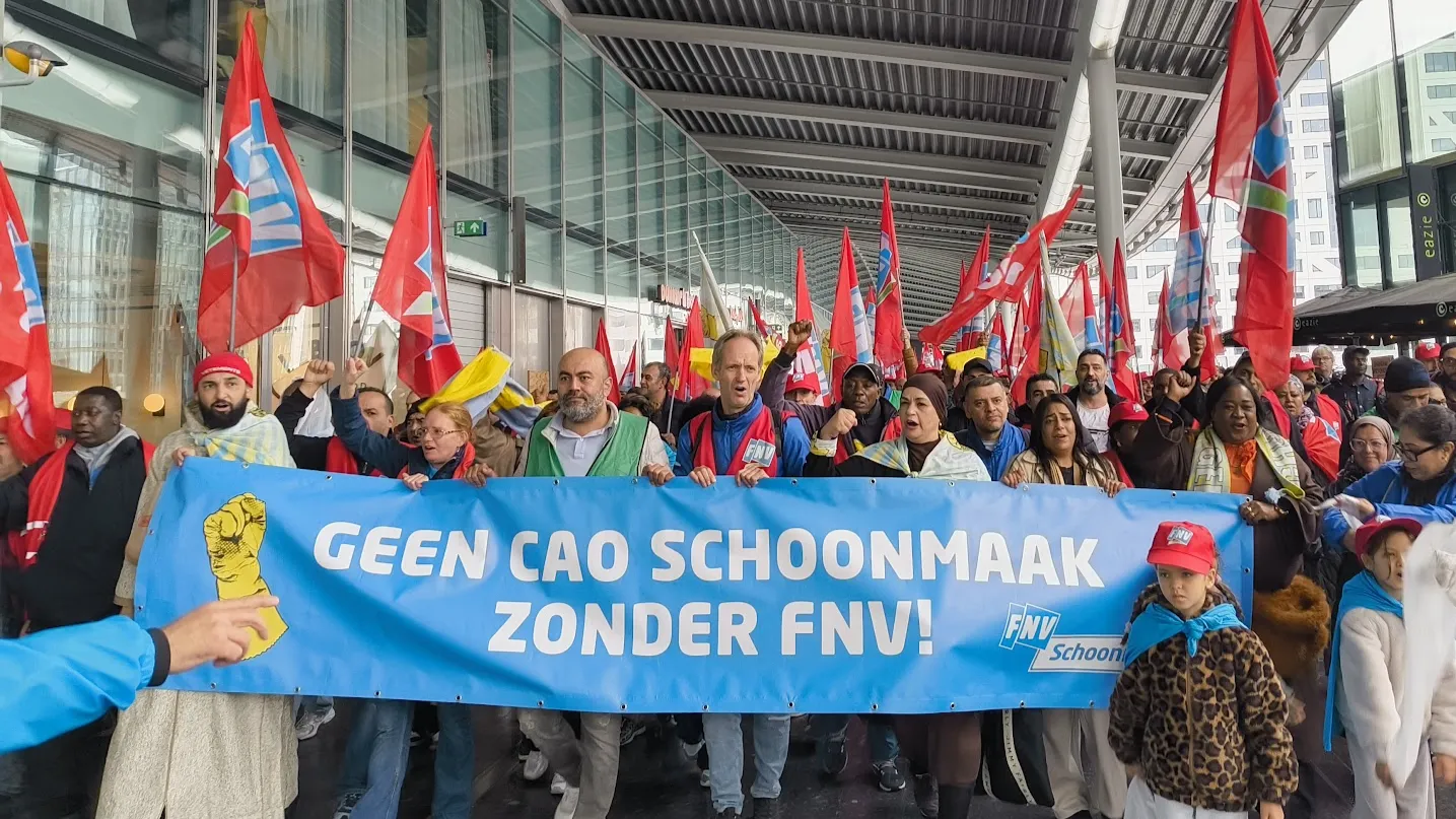 Stakende schoonmakers lopen langs Utrecht Centraal. Op het spandoek staat: "Geen cao schoonmaak zonder FNV!"