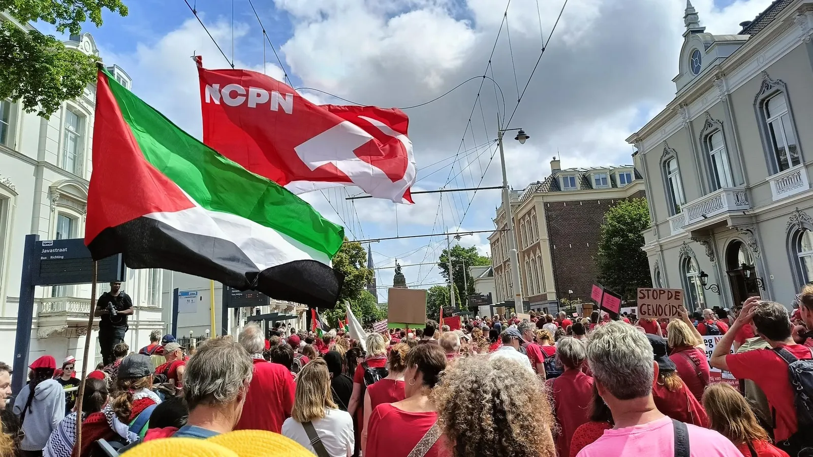 Een enorme groep rood geklede mensen loopt door de straten in Den Haag, met op de voorgrond een wapperende Palestijnse vlag en NCPN-vlag