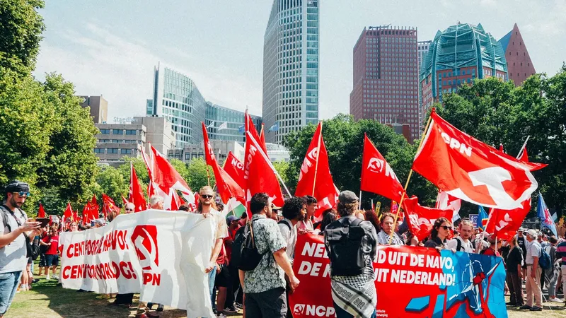 Mensenmassa met spandoeken en rode vlaggen tijdens protest tegen de NAVO-top in Den Haag op 22 juni, met NCPN en CJB zichtbaar vooraan.