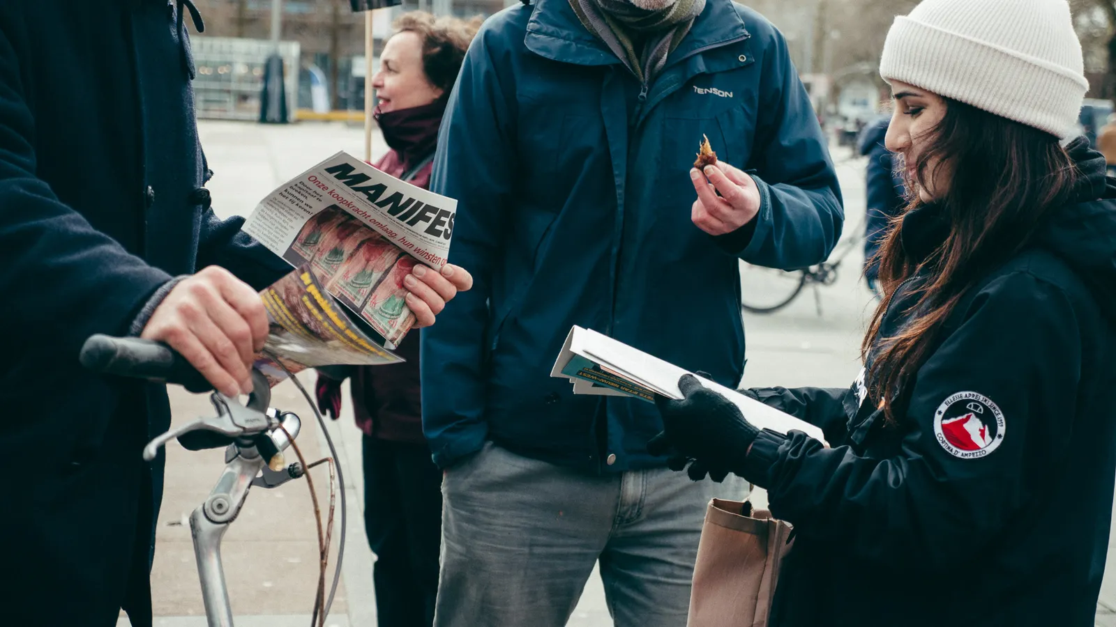 Iemand geeft deelt een Manifest-editie uit aan voorbijgangers op een plein, die aandachtig naar haar staan te luisteren
